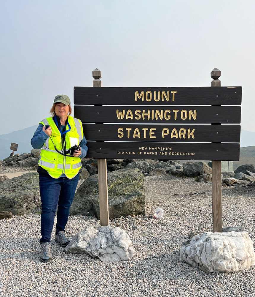 Deb Camejo in a yellow vest and cap smiles, standing beside a "Mount Washington State Park" sign.