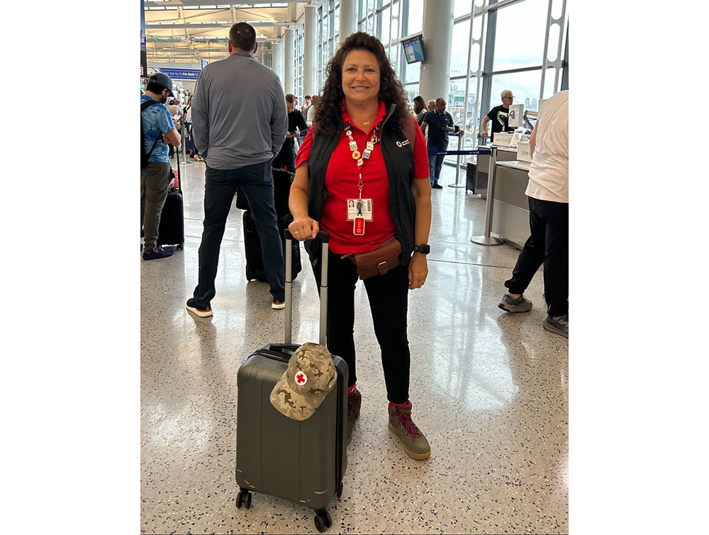 Dee Chase in airport terminal with Red Cross hat on top of rolling luggage, wearing Red Cross shirt and vest.