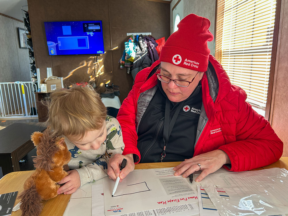 Red Cross volunteer showing fire escape route to child at a kitchen table.