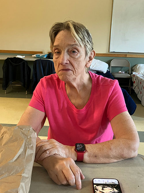 Woman sitting at table with brown paper bag and cell phone, wearing a watch looking at camera.