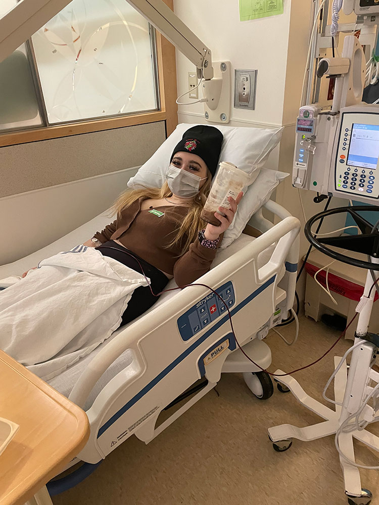 Jade Howard, wearing a beanie and mask, lying in hospital bed getting a blood transfusion.
