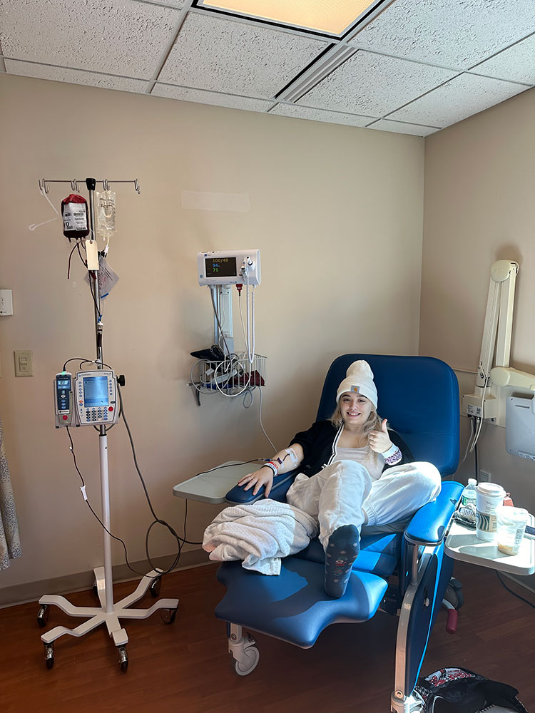 Jade Howard giving a thumbs up and sitting in hospital chair getting a blood transfusion.
