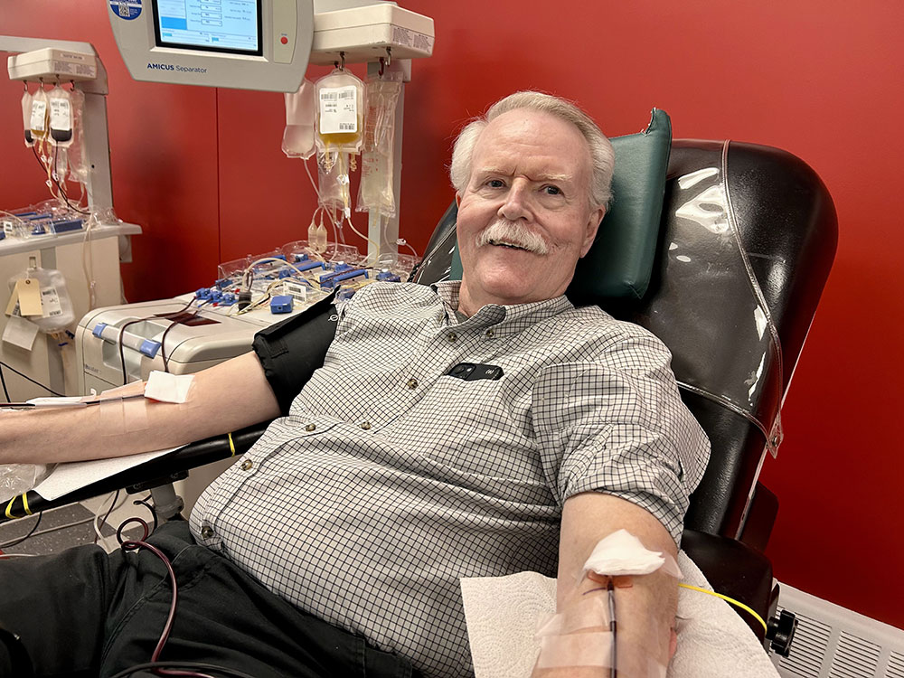 John Burke in a plaid shirt smiles while donating blood, seated in a dark chair. Medical equipment with tubes and bags is visible against a red wall.