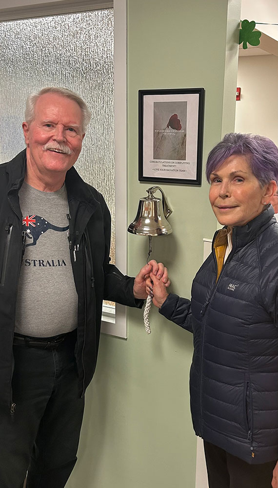 John and Christine Burke stand smiling beside a silver bell on a wall. They hold the bell's rope together.