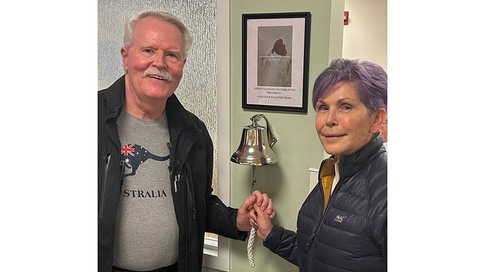 John and Christine Burke stand smiling beside a silver bell on a wall. They hold the bell's rope together.