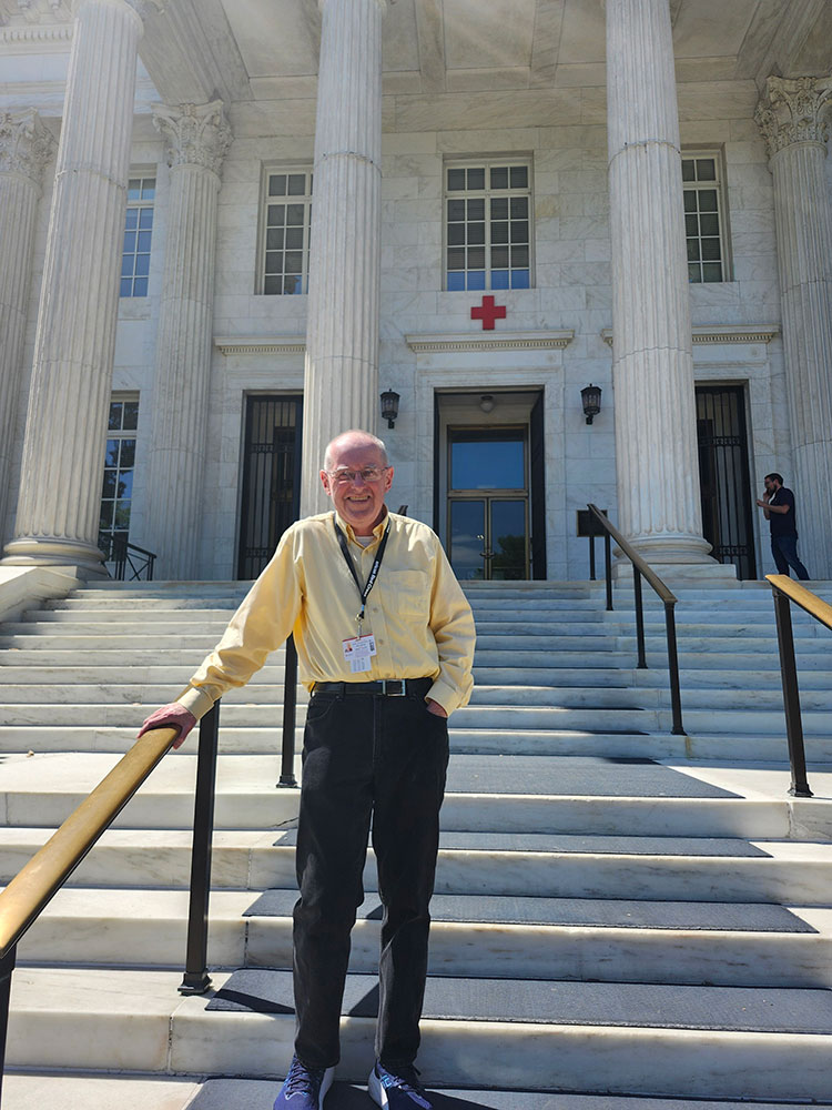 John Leeming in a yellow shirt stands smiling on the steps of a grand building with tall columns and a red cross symbol.