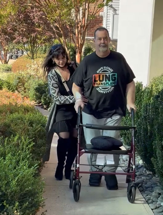 Amy Deavitt helping Keith Deavitt walk with his walker on a sidewalk.