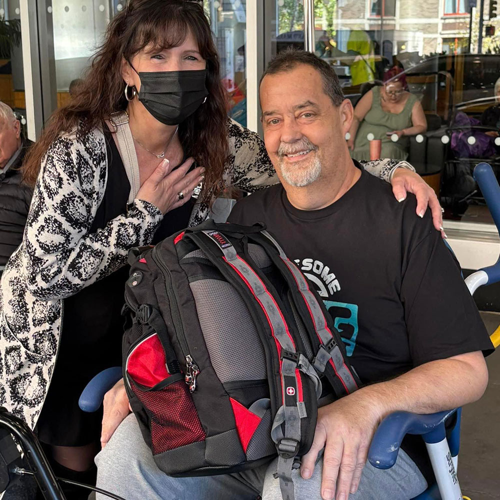 Amy Deavitt, wearing a mask, standing next to Keith Deavitt who is sitting in a chair outside.