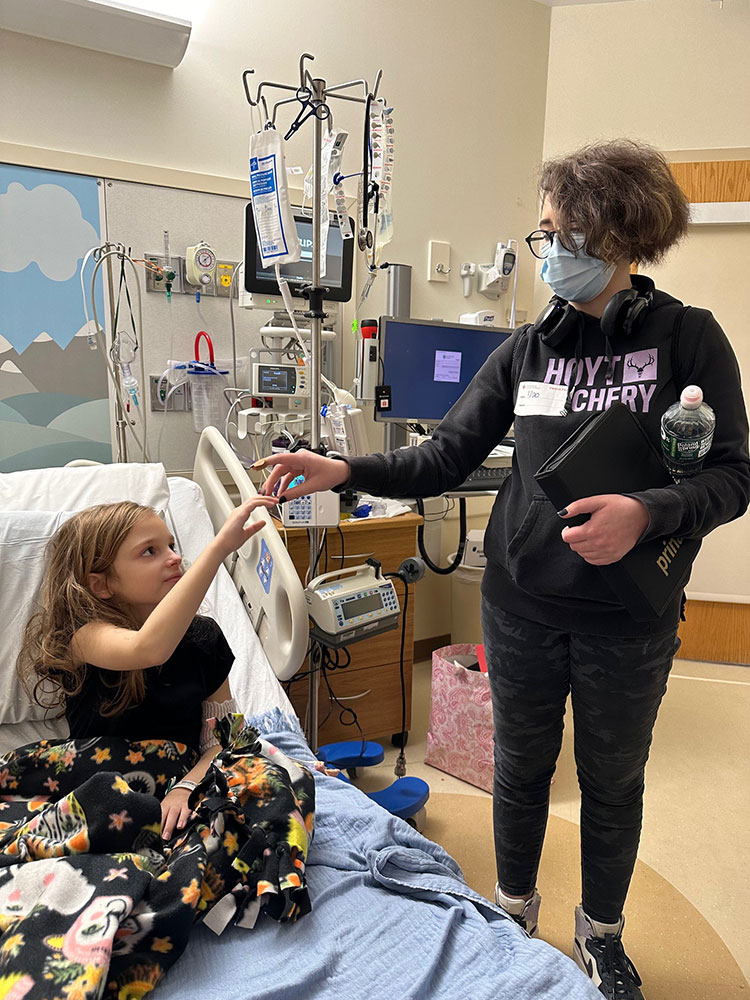 Lucy Bellemare in hospital bed touching the hand of a person standing next to her.