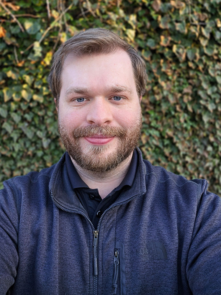 Max Knecht with a beard smiling in front of leafy greenery. He wears a dark jacket.