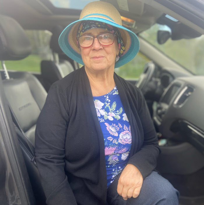 An older woman sits in a car wearing glasses, a scarf around her head and a hat.
