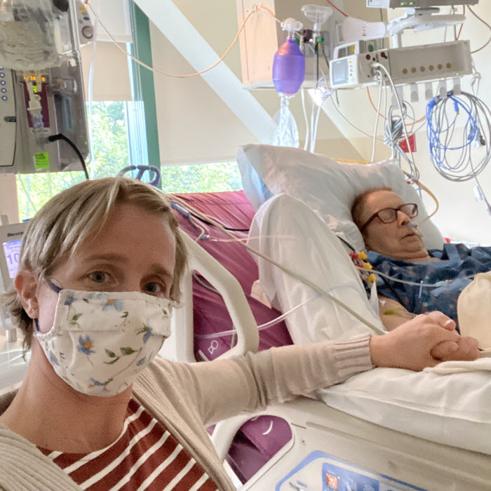 An older woman laying in a hospital bed with her eyes closed while her younger daughter, wearing a mask, holds her hand.