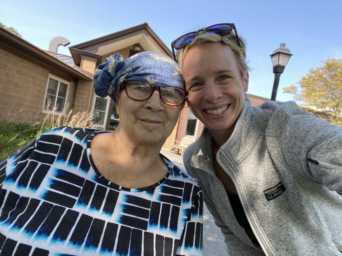 An older woman wearing a scarf around her head and glasses stands outside next to her yournger daughter. Both are smiling.