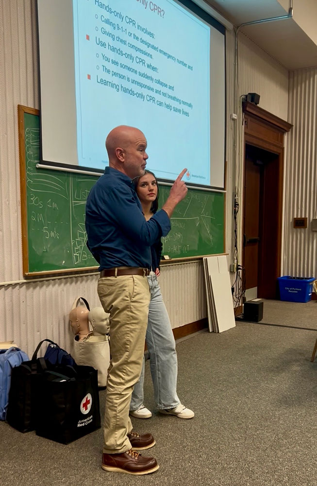 Peter Morrison and Kate Morrison stand together in front of a classroom, engaged in conversation with students in the background.