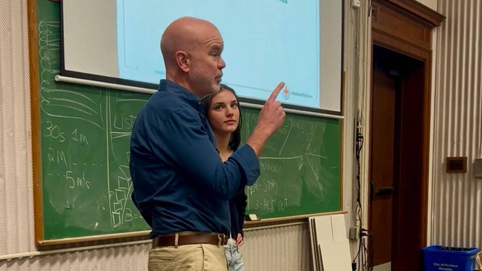 Peter Morrison and Kate Morrison stand together in front of a classroom, engaged in conversation with students in the background.