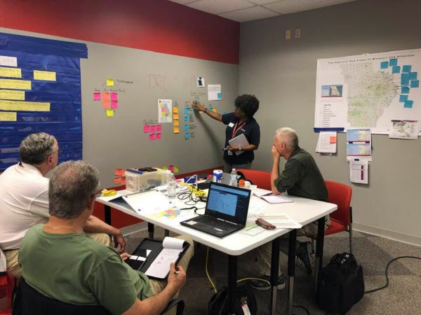 Phyllis stands at a wall covered in post in notes writing something while 3 men sitting at a table look on