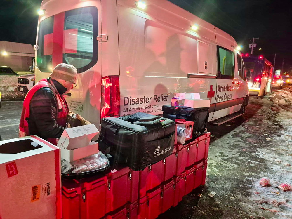 Red Cross volunteer, wearing a beanie and jacket, standing next to Red Cross boxes on the side of a snowy road and a Red Cross van.
