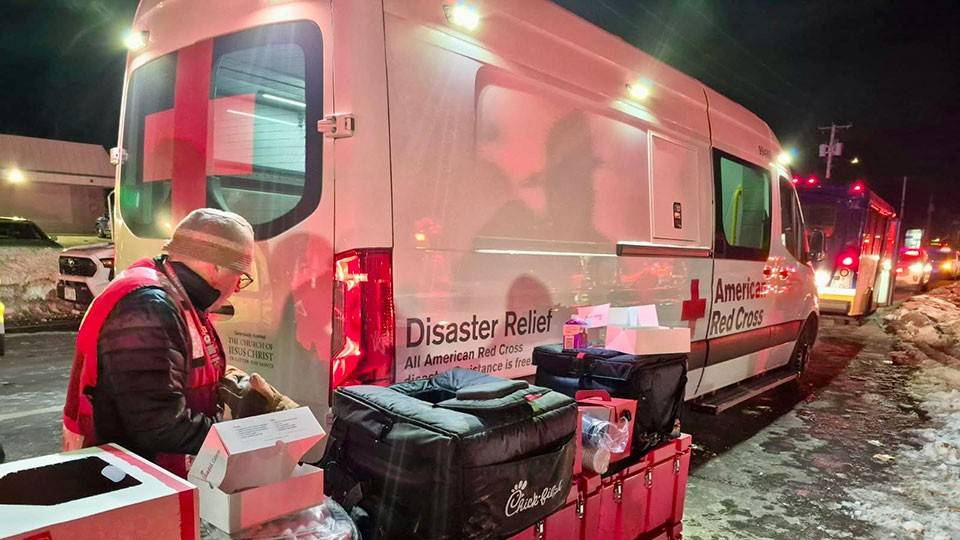 Red Cross volunteer, wearing a beanie and jacket, standing next to Red Cross boxes on the side of a snowy road and a Red Cross van.