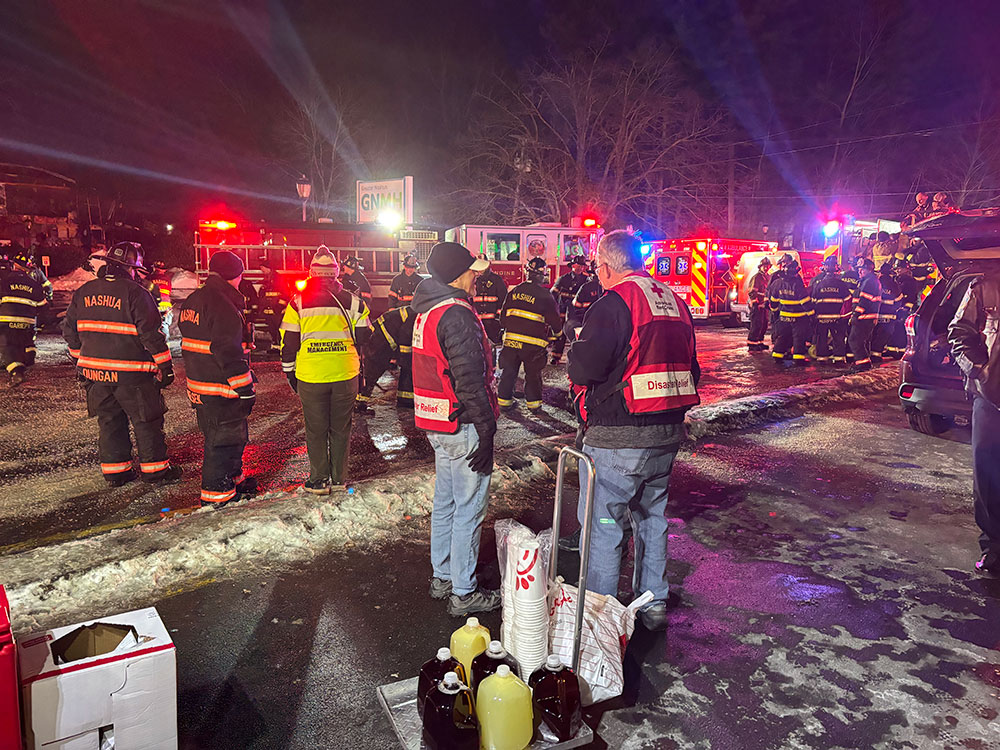 Red Cross volunteers and firefighters standing by fire engines on a snowy road.