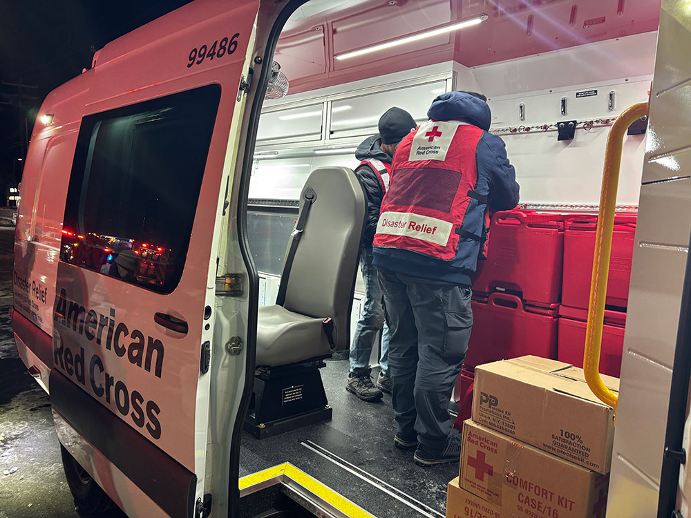 Red Cross volunteers unloading supplies from a Red Cross van on a snowy road.