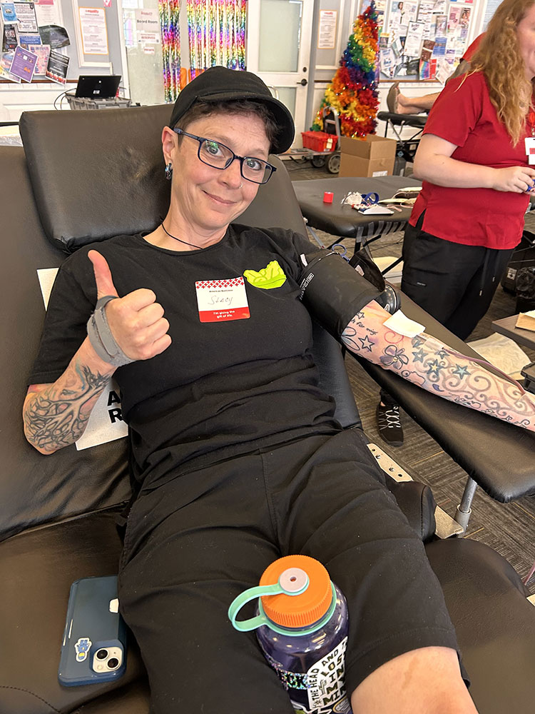 Stacy Nicols sitting in a blood donation chair, giving a thumbs up, and donating blood.