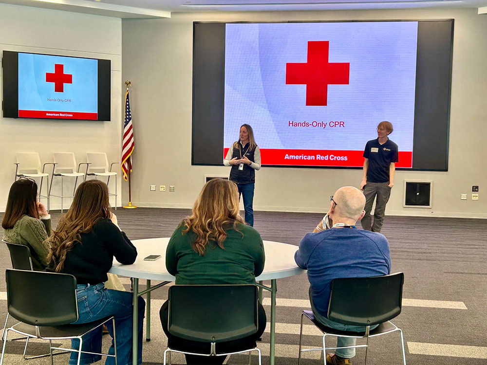 Group seated at a training session led by two Red Cross instructors at the front, with a large screen displaying "Hands-Only CPR" and American Red Cross logo.