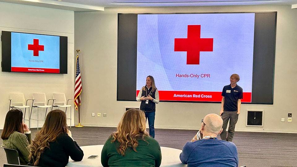 Group seated at a training session led by two Red Cross instructors at the front, with a large screen displaying "Hands-Only CPR" and American Red Cross logo.