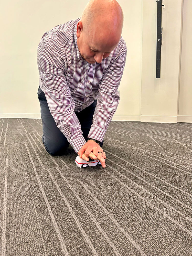 A man in a checkered shirt kneels on a carpeted floor, practicing CPR compressions on a small training device.