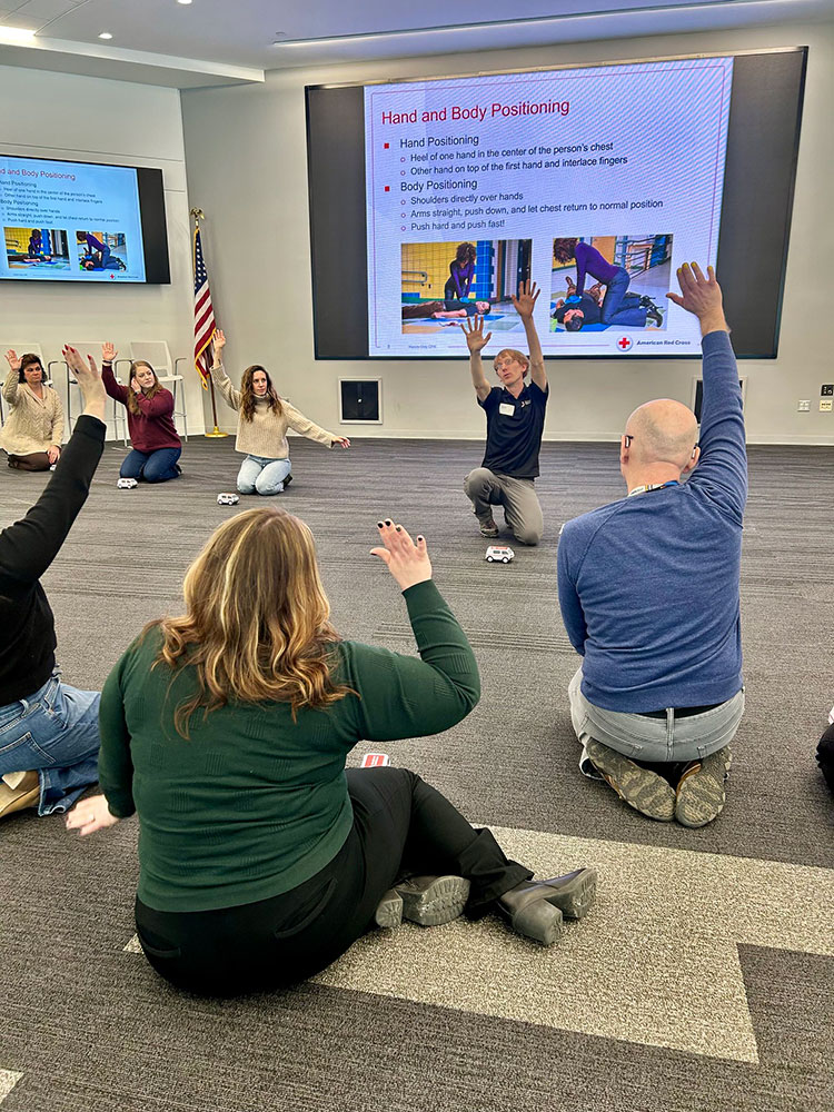 Group CPR training session with people kneeling on the floor, raising hands. Large screen displays instructions for hand and body positioning.