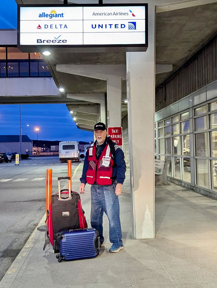 Red Cross volunteer Paul Miller waits with his luggage by the front doors of an airport.