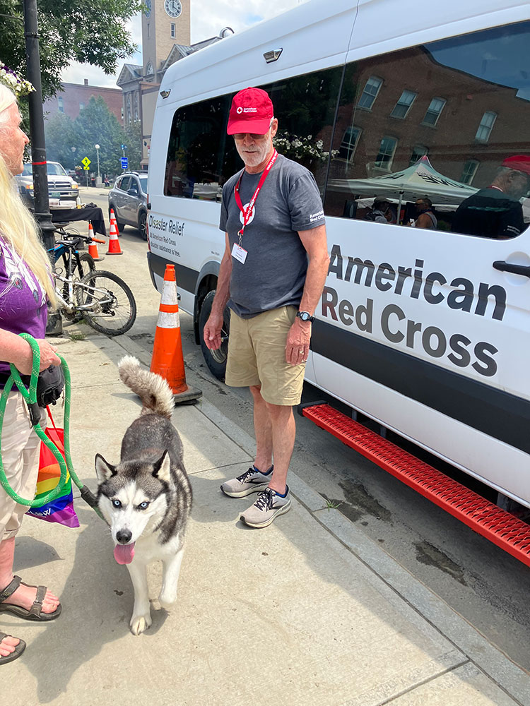 Red Cross volunteer Pete Basiliere standing by a Red Cross vehicle and looking at a dog on a leash on the sidewalk.