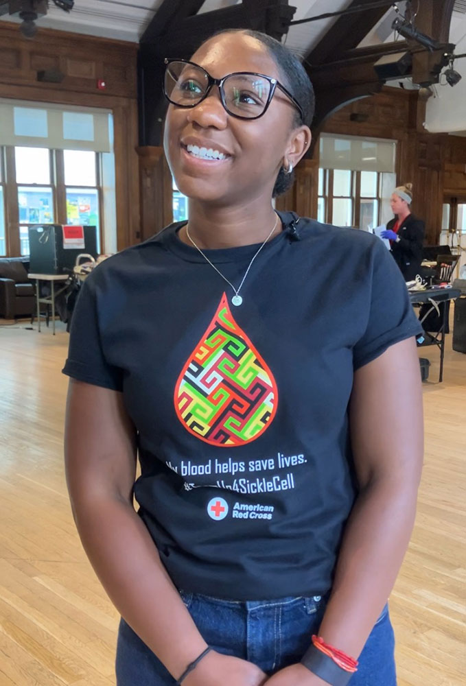 college student wearing glasses and a Red Cross shirt at the blood drive inside a room where people are donating blood.