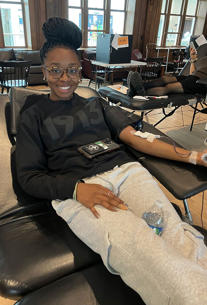 college student sitting in chair with smartphone on lap, donating blood at the blood drive.