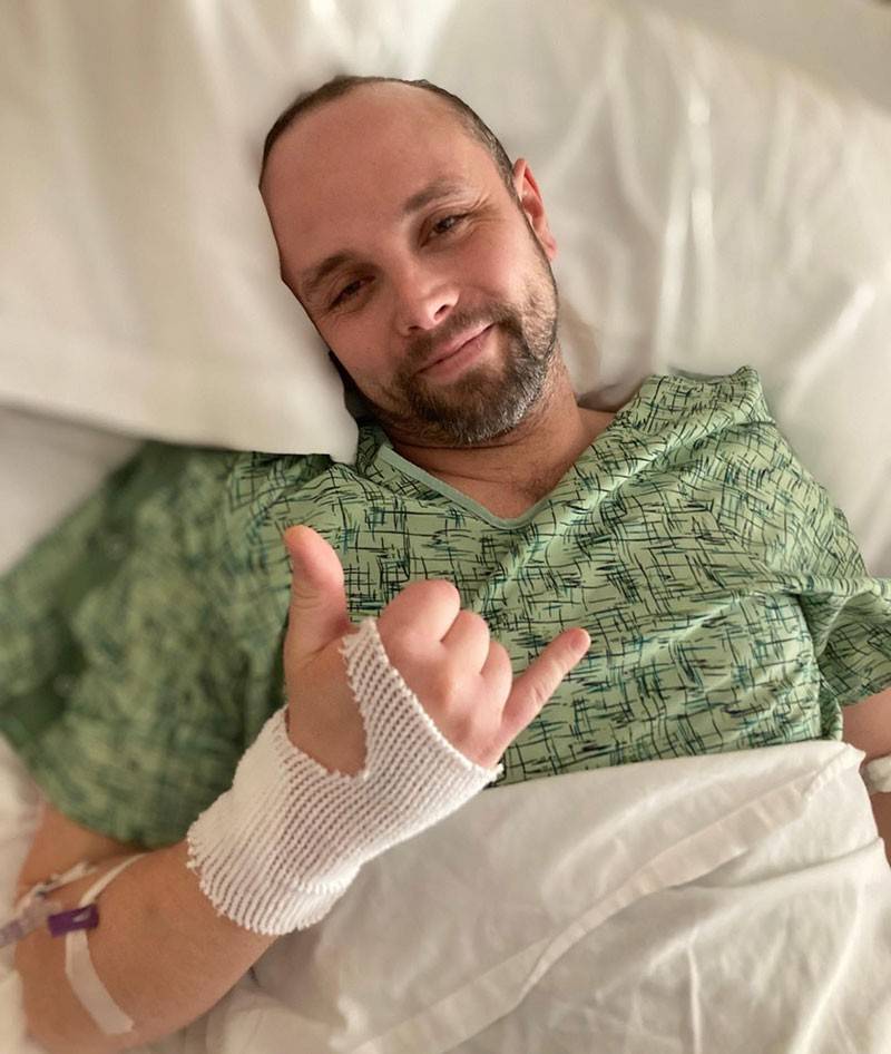Alex Adams in hospital bed giving hang loose sign