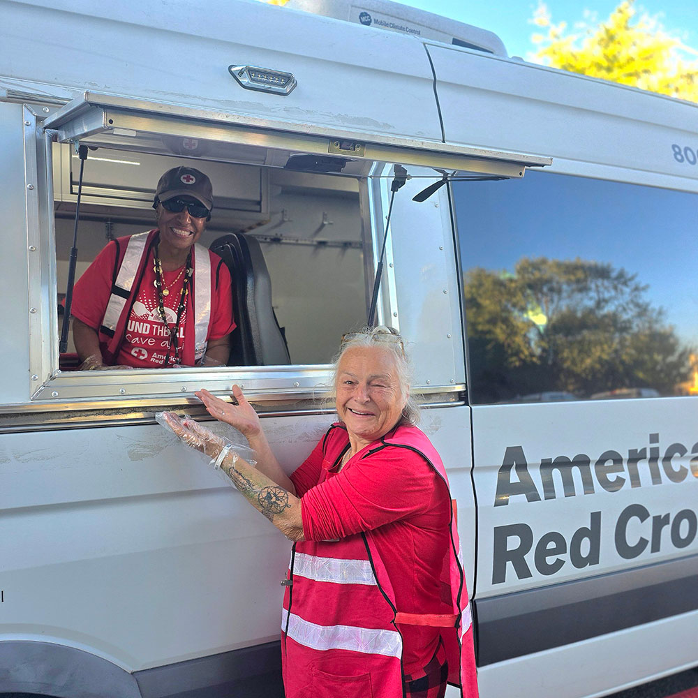 Two Red Cross volunteers with one in a Red Cross vehicle and the other standing outside of the vehicle.