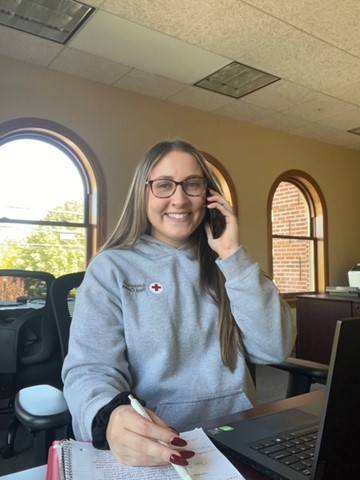 Emma Banton wearing glasses and red cross hoodie, talking on phone while sitting at a desk with a laptop.