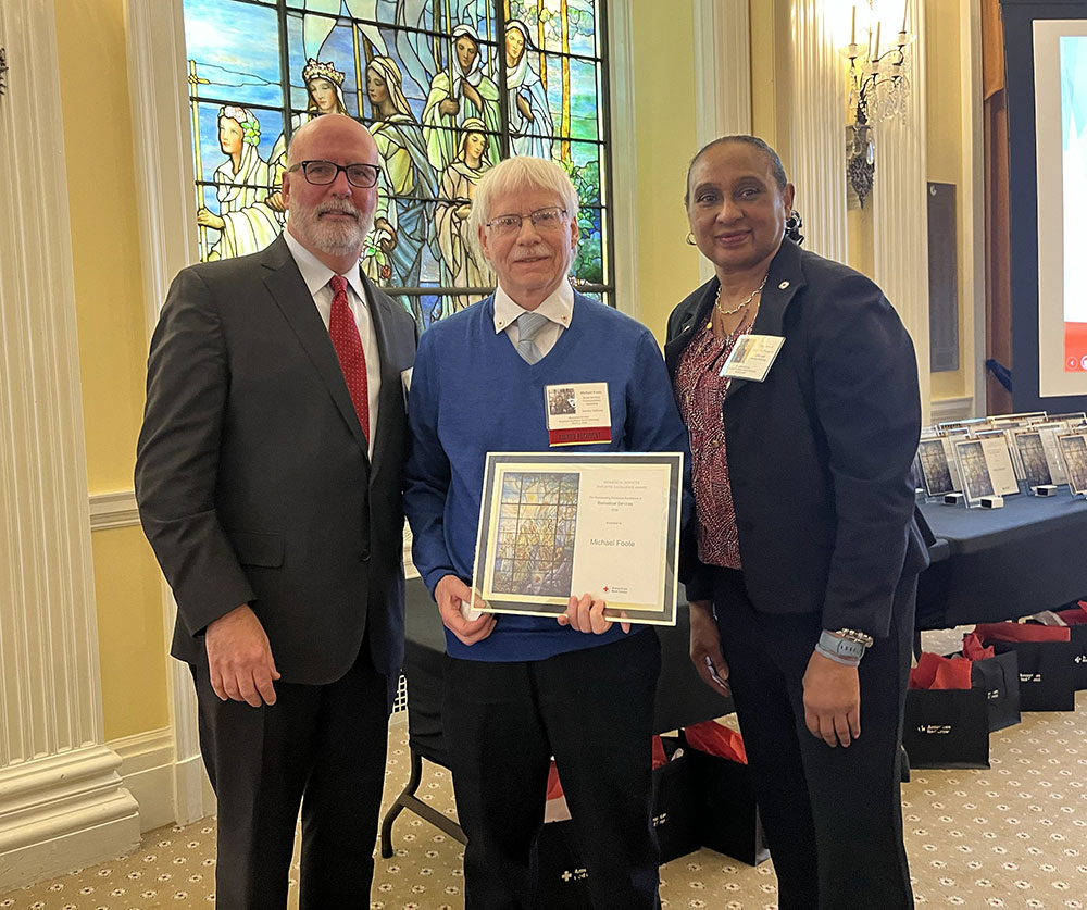 Three people stand together smiling. Michael Foote holds a certificate. Stained glass and awards are visible in the background, creating a formal atmosphere.