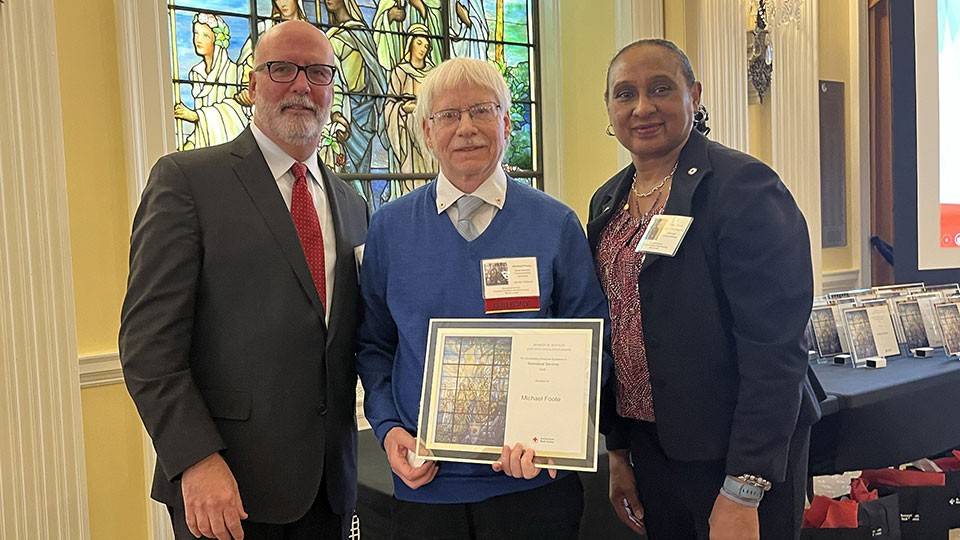 Three people stand together smiling. Michael Foote holds a certificate. Stained glass and awards are visible in the background, creating a formal atmosphere.