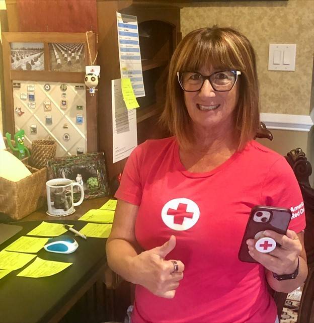 Monica Bunner wearing glasses and red cross shirt and holding a smart phone and giving a thumbs up while sitting a desk.