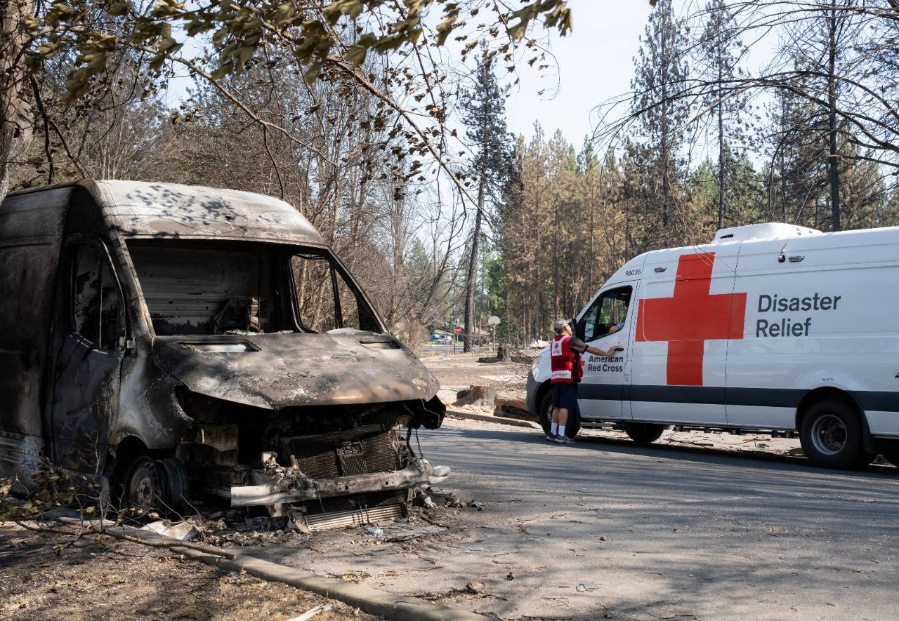 A man in a red cross vest stands outside a van next to a burned vehicle