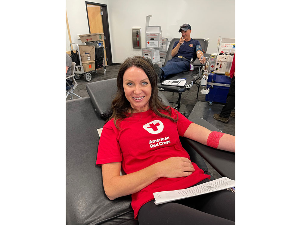 Person wearing an American Red Cross shirt sitting in cot donating blood and smiling for camera.