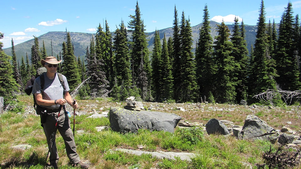 Bob Helmer wearing hat, sunglasses, backpack and holding walking sticks in a wooded area.