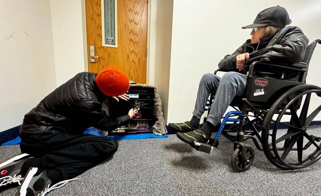 Woman sits in wheelchair as man checks on dog in kennel