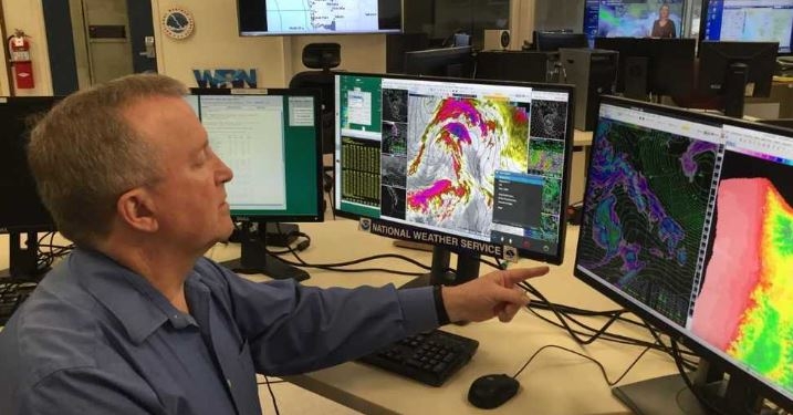 Man sits at desk looking at computer screens showing weather patterns