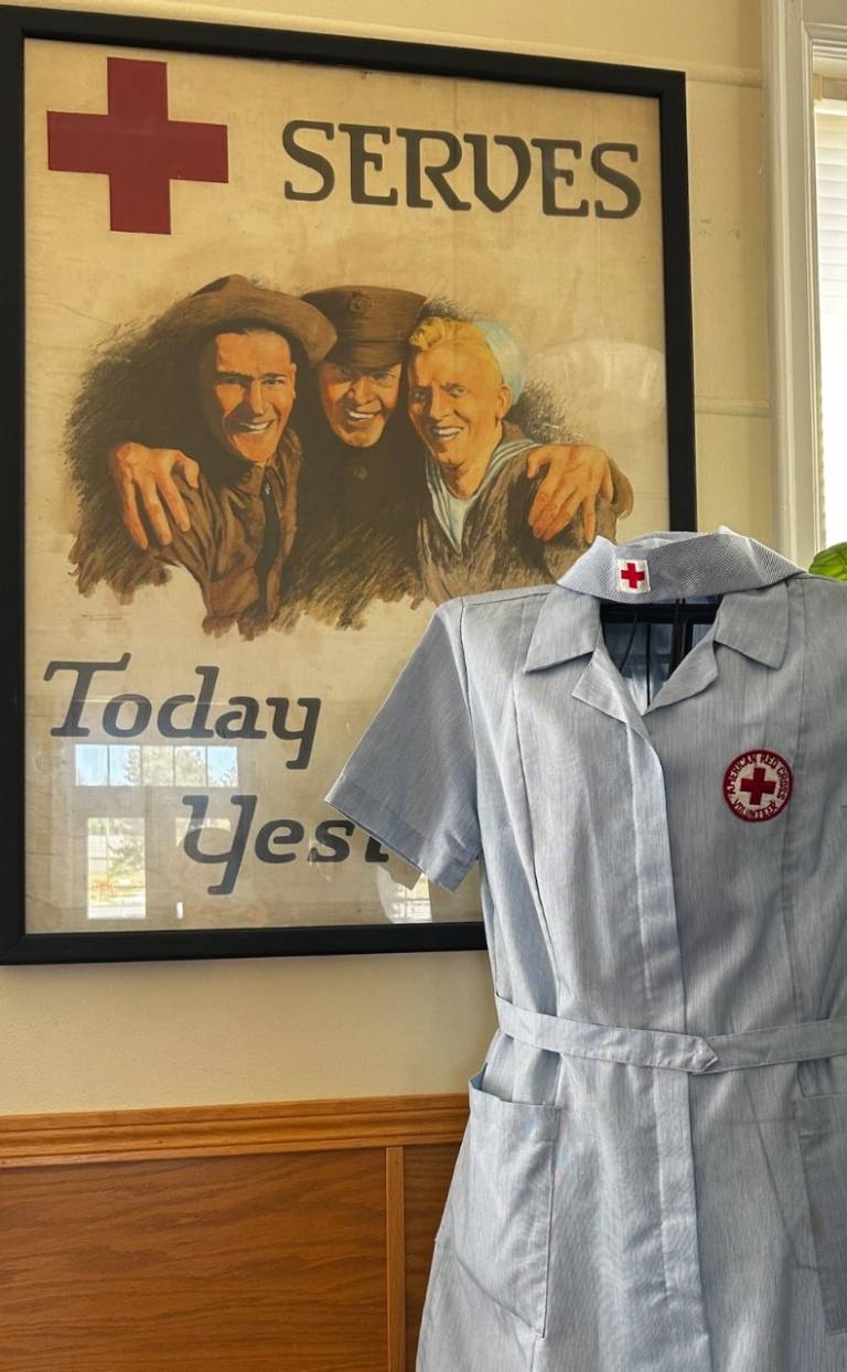 A vintage Red Cross uniform hanging on a dress form in front of a World War 2 era poster