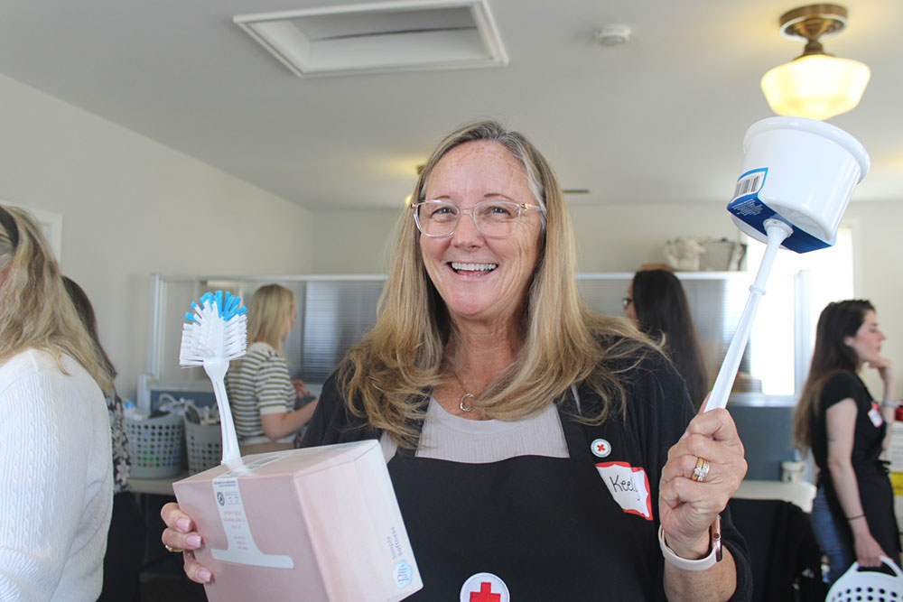 Woman with glasses and Red Cross apron holds new cleaning supplies and smiling for the camera.