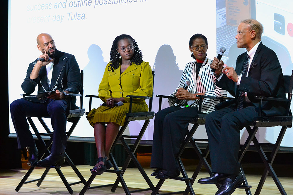 four people holding mics while sitting in tall folding chairs on a stage.