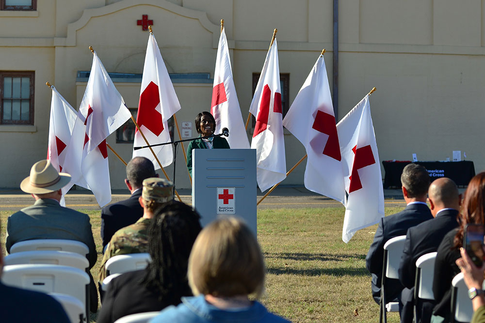 a person speaking at a podium to a seated audience on the grass with Red Cross flags and building in the background.