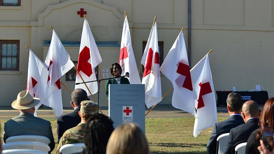 a person speaking at a podium to a seated audience on the grass with Red Cross flags and building in the background.