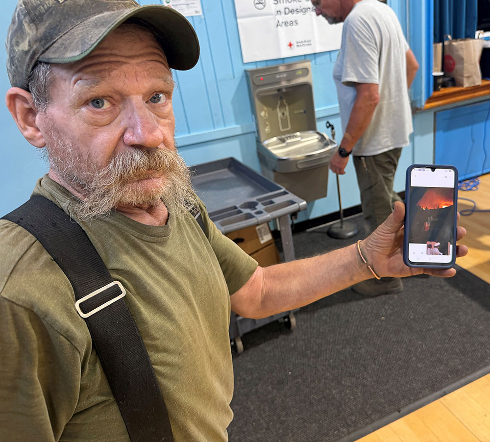 Tim Webb wearing a camo hat and holding a smart phone that has a picture of a forest fire.